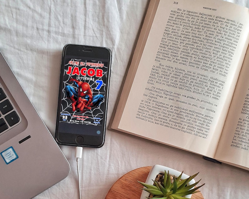 Peut inclure: Un smartphone noir affichant une invitation d'anniversaire Spiderman. L'invitation est pour le 7&egrave;me anniversaire de Jacob et pr&eacute;sente une illustration de Spiderman rouge et bleu. Le t&eacute;l&eacute;phone est connect&eacute; &agrave; un c&acirc;ble de charge blanc.