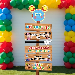 May include: A colorful party decoration featuring a balloon arch in red, yellow, green, and blue. A wooden sign with the text "OH TOODLES", "THIS WAY TO", "SEBASTIAN'S 1ST BIRTHDAY", "COME!! ITS FUN INSIDE", and "LETS PARTY" is displayed. Mickey and Minnie Mouse are featured.