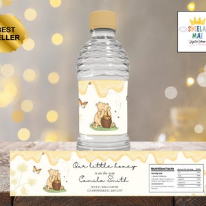 Puede incluir: Una botella de agua transparente con tapa amarilla y una etiqueta con un oso de dibujos animados y un diseño de goteo de miel. La etiqueta dice "Our little honey is on the way Camila Smith". Una pegatina de "Best Seller" es visible.