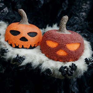 Puede incluir: Dos calabazas de Halloween de ganchillo, una naranja y otra burdeos, con caras talladas. Las calabazas están sobre una superficie blanca y esponjosa sobre un fondo oscuro. La calabaza naranja tiene una cara negra y la burdeos tiene un brillo naranja.