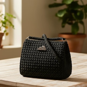 Può includere: Una borsa nera con una trama intrecciata, con un manico superiore e una chiusura argentata. La borsa è appoggiata su un tavolo di legno chiaro. La chiusura riporta il nome del marchio.