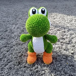 Könnte beinhalten: Eine handgefertigte grüne und weiße Yoshi-Puppe aus Häkelarbeit mit orangefarbenen Stiefeln. Das Plüschtier hat große schwarze und weiße Augen und einen runden Kopf. Die Puppe steht auf einem grauen Teppich.