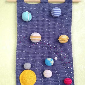 May include: A crocheted wall hanging depicting a solar system with planets of different colours and patterns, including a yellow sun, a blue and white striped planet, a pink and white striped planet, a brown and white striped planet, a blue planet with a white swirl, a grey planet, and a red planet. The planets are surrounded by white and silver beads, representing stars.
