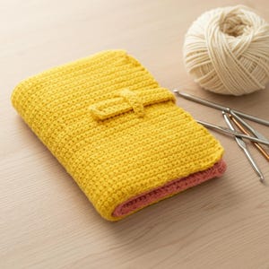 Puede incluir: Una funda amarilla tejida a ganchillo con cierre de correa a juego. La funda está doblada, revelando un interior rosa. Una madeja de hilo color crema y varios ganchillos están junto a la funda, sugiriendo un tema artesanal.