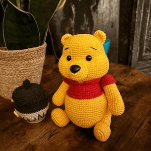 Könnte beinhalten: Ein handgefertigter gelber Häkelbär mit rotem Shirt. Der Bär hat schwarze Augen und eine schwarze Nase. Neben dem Bären befindet sich ein schwarz-weißes Häkelobjekt. Der Bär sitzt auf einer Holzoberfläche.