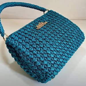 Puede incluir: Un bolso de mano de crochet azul turquesa con un cierre de metal dorado. El bolso tiene una sola correa y una textura tejida.