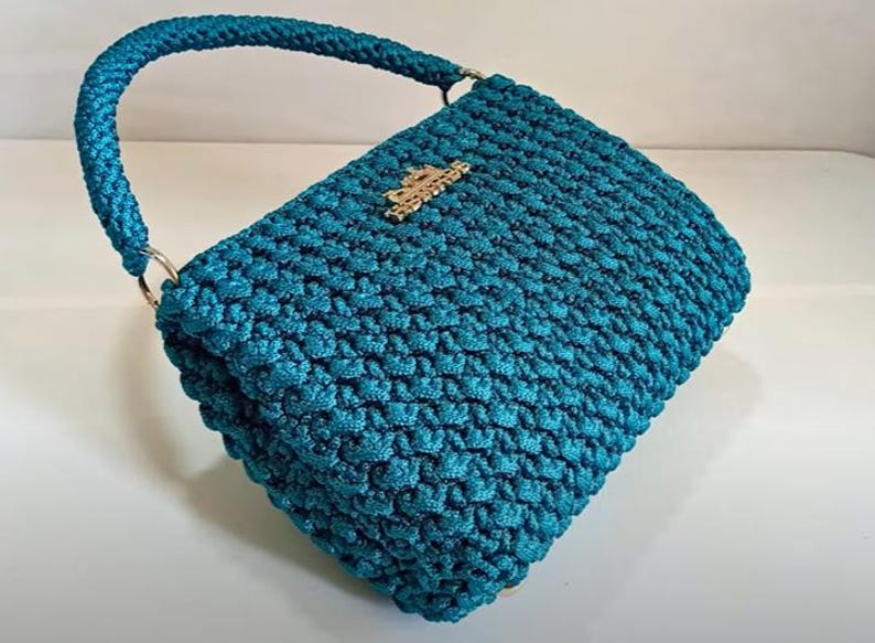 Puede incluir: Un bolso de mano de crochet azul turquesa con un cierre de metal dorado. El bolso tiene una sola correa y una textura tejida.