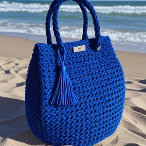 以下が含まれることがあります： 明るい青色のクロシェのトートバッグと、それに合わせたタッセル。バッグにはロープのハンドルと、「JJ」という文字が書かれた銀色のタグが付いています。バッグは砂浜の上に置かれています。
