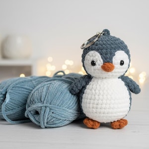 May include: A crocheted penguin keyring with a white belly, grey head and wings, and orange beak and feet. The penguin is next to three balls of blue yarn. The background is blurred with soft lighting.
