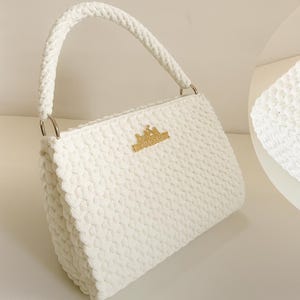 Pode incluir: Bolsa branca de crochê com textura e alça a condizer. Apresenta argolas prateadas e um emblema dourado. A bolsa tem formato retangular.
