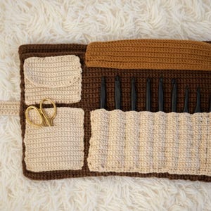 May include: A brown and beige crochet hook organizer with multiple pockets. The organizer holds black crochet hooks and gold scissors. The organizer is made of brown and beige crochet yarn.