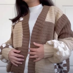 May include: A brown and beige crocheted cardigan with a patchwork design. The cardigan has long sleeves and a v-neck.