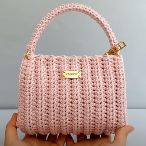 May include: A light pink crochet handbag with a handle and a gold-colored zipper. The bag has a rectangular shape and a small gold-colored tag that reads "FASHION". The bag is held by a person's hands.