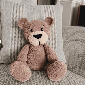 May include: A brown crocheted teddy bear with black eyes and a black nose. The bear is sitting on a gray and white striped pillow.