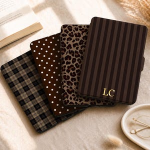May include: Four rectangular e-reader cases arranged in a fan shape. The patterns include a black and tan plaid, a dark brown with white polka dots, a brown and black leopard print, and a dark brown vertical striped case with the gold letters LC.