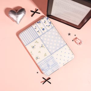 May include: A rectangular e-reader case featuring a patchwork design of light blue and white patterns, including stripes, polka dots, and small floral motifs. The case is surrounded by a silver heart, black ribbon bows, and small star-shaped confetti.