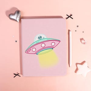 Op de afbeelding: Een roze tablethoes met een cartoon UFO en alien ontwerp. De UFO is roze, groen en blauw, met een gele lichtstraal. Een witte stylus ligt ernaast. De achtergrond is roze met sterren en decoratieve vormen.