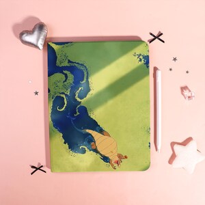 Op de afbeelding: Een groen notitieboek met een blauw en groen illustratie van een wezen. Een witte stylus en decoratieve items liggen naast het notitieboek op een roze ondergrond.