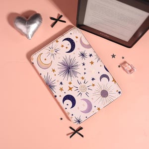 May include: A light pink Kindle cover with a celestial design featuring moons, stars, and suns in navy blue, gold, and purple. A silver heart-shaped pillow and a Kindle e-reader are also visible.