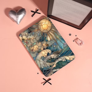May include: A Kindle case with a stained-glass design depicting a sun, clouds, and ocean waves in blue, gold, and white. The case is on a pink surface, next to a silver heart-shaped object and a Kindle e-reader.
