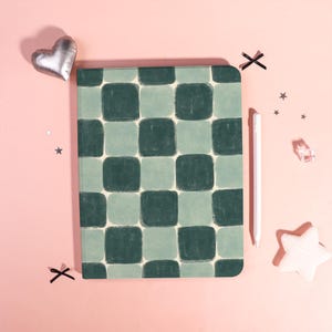May include: A notebook with a green and light green checkerboard pattern. A white stylus, a silver heart-shaped decoration, and a white star-shaped decoration are next to the notebook on a pink surface.