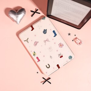 May include: A light pink e-reader case featuring a scattered pattern of small illustrations including cowboy boots, hats, a bull skull, bows, flowers, cherries, a horseshoe, and stars. The case is positioned next to a black Kindle e-reader.