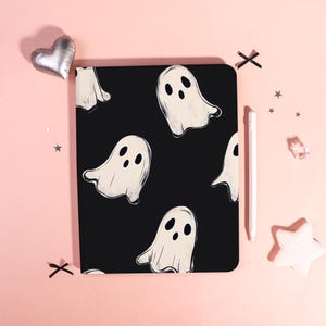 May include: Black notebook with a repeating pattern of white ghost illustrations. The notebook is surrounded by decorative elements, including a silver heart-shaped pillow, star-shaped confetti, and a white stylus.