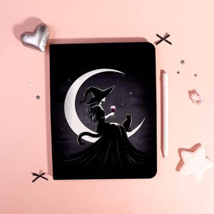 Pode incluir: Caderno preto com uma ilustração de uma bruxa sentada numa lua crescente, segurando um copo de vinho, com um gato preto. A bruxa usa um chapéu pontudo e um vestido longo preto contra um céu estrelado.