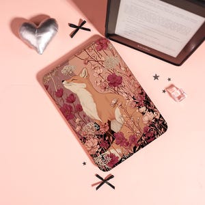 Peut inclure: Une housse de liseuse rectangulaire ornée d'une illustration imprimée représentant un renard entouré de fleurs roses et rouge foncé. L'étui présente une finition lisse et est présenté avec un objet argenté en forme de cœur et des nœuds noirs.