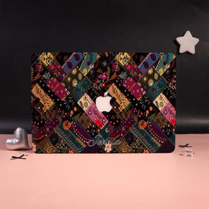 Op de afbeelding: Een laptopskin met een diagonaal patchworkpatroon van donkere, veelkleurige bloemmotieven en geometrische ontwerpen. Het oppervlak toont een verscheidenheid aan kleine, ingewikkelde motieven in tinten rood, blauw, geel en groen. De naam Jennia staat in wit schrift bij het midden, onder de uitsparing voor het Apple-logo.