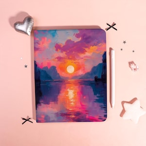 Könnte beinhalten: Ein Notizbuch mit einem lebendigen Sonnenuntergangs-Landschaftsdesign. Das Cover zeigt ein farbenfrohes Gemälde eines Sonnenuntergangs über dem Wasser, mit Farbtönen von Rosa, Orange und Lila. Ein weißer Stift und dekorative Gegenstände befinden sich neben dem Notizbuch.