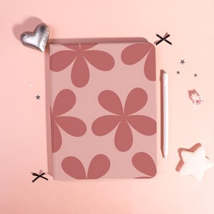 Peut inclure: Une coque de tablette rose avec un motif floral rose foncé. Un stylet blanc, un ballon argenté en forme de cœur et des confettis en forme d'étoile sont disposés autour de la coque sur une surface rose.