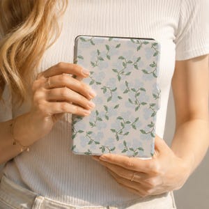 May include: A rectangular e-reader case featuring a repeating pattern of light blue hydrangea clusters and green leaves on a white background. The case has a smooth, flat surface and a distinct side tab for opening, showcasing a floral botanical design.