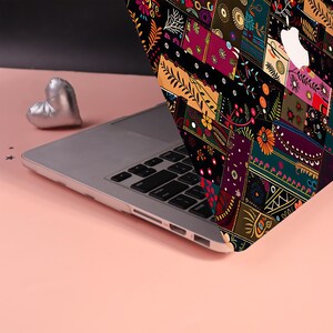 Może przedstawiać: Laptop z etui ochronnym ozdobionym żywym, wielokolorowym wzorem patchworkowym z motywami kwiatowymi i geometrycznymi. Laptop posiada srebrną klawiaturę oraz białe logo jabłka na pokrywie. Obok znajduje się mały, srebrny przedmiot w kształcie serca.