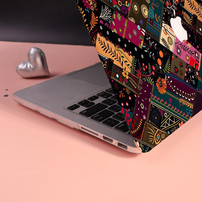 Może przedstawiać: Laptop z etui ochronnym ozdobionym żywym, wielokolorowym wzorem patchworkowym z motywami kwiatowymi i geometrycznymi. Laptop posiada srebrną klawiaturę oraz białe logo jabłka na pokrywie. Obok znajduje się mały, srebrny przedmiot w kształcie serca.