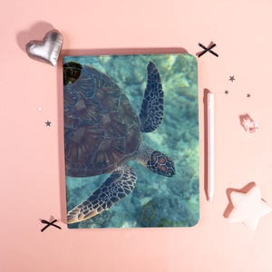 May include: A notebook featuring a detailed image of a sea turtle swimming underwater. The turtle has a brown shell with a patterned design, and its flippers are extended. The background is a light blue, creating a serene aquatic scene.