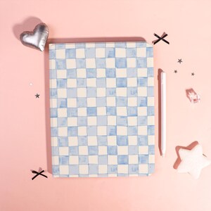 May include: A notebook with a blue and white checkered pattern. A white stylus rests beside the notebook on a pink surface. Decorative elements include a silver heart, star-shaped confetti, and a white star.