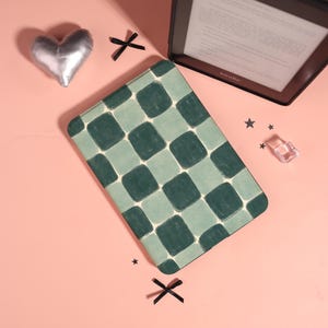 Może przedstawiać: Zielone i jasnozielone etui na Kindle w kratkę spoczywa na różowej powierzchni, obok czytnika e-booków Kindle. Srebrna poduszka w kształcie serca, czarna kokarda i małe gwiazdki również znajdują się na powierzchni. Etui jest prostokątne i ma powtarzający się kwadratowy wzór.
