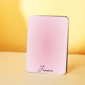 Puede incluir: Un espejo compacto rectangular rosa con un borde negro. El nombre "Jennia" está escrito en cursiva en la parte inferior. El fondo es un degradado de amarillo y rosa.