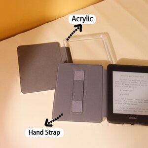 May include: A gray e-reader case with a clear acrylic cover and a hand strap. The case is closed and the e-reader is visible inside. The e-reader screen displays text in French.