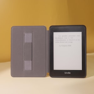 May include: A black Kindle e-reader with a gray case. The case has a built-in hand strap. The screen displays text in French, "Les grandes personnes sont d&eacute;cid&eacute;ment tout &agrave; fait extraordinaires, se disait-il en lui m&ecirc;me durant son voyage. Le Chapitre XIII."