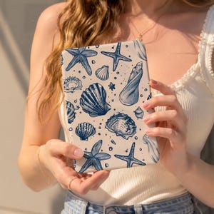 Può includere: Una custodia rettangolare per e-reader con sfondo color crema e un motivo ripetuto di illustrazioni blu scuro di varie conchiglie e stelle marine, tra cui conchiglie a spirale, capesante e stelle marine a cinque punte sparse.