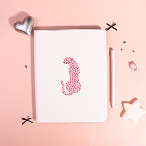 Può includere: Un quaderno bianco con un disegno di ghepardo rosa fatto di stelle. Uno stilo bianco, un cuore argentato, una stella bianca e forme a X nere sono sparse su uno sfondo rosa. Il quaderno è rettangolare.