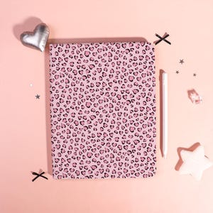 Può includere: Un taccuino rosa con un motivo leopardato a forma di cuore. Uno stilo bianco, una decorazione a forma di cuore argentata e coriandoli a forma di stella sono disposti attorno al taccuino su uno sfondo rosa.