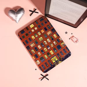 May include: A rectangular e-reader case featuring a detailed illustration of a multi-story brick apartment building at night. The facade shows numerous illuminated windows with various figures, holiday decorations, and interior scenes depicted in a grid.