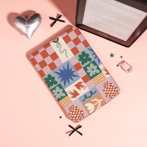 Peut inclure: Une housse de liseuse rectangulaire avec un motif à carreaux multicolores composé de carrés terracotta, lavande et crème. Le design présente des illustrations de fleurs vertes, une étoile bleue, deux oiseaux bruns et des formes abstraites.