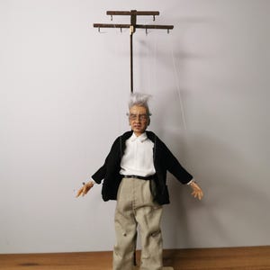 Puede incluir: Una marioneta que representa a una persona mayor con el pelo gris, vestida con una camisa blanca de cuello, una chaqueta negra y pantalones beige. La figura está suspendida por hilos blancos finos de una barra de control de madera en cruz.