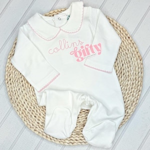 May include: White baby sleeper with a Peter Pan collar and pink trim. The sleeper features the embroidered name "collins" and the word "gifty" in a pink, stylized font. The sleeper is made of soft material.