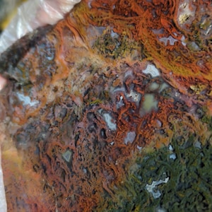 Puede incluir: Primer plano de una piedra de ágata colorida con patrones rojos, naranjas, amarillos, verdes y blancos. La piedra tiene una textura rugosa y natural.