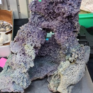May include: A large grape agate specimen, showcasing a cluster of rounded, purple formations. The mineral exhibits a range of violet shades, with a bubbly texture. The agate sits on a silver scale, with a pink label that reads "Grape Agate".
